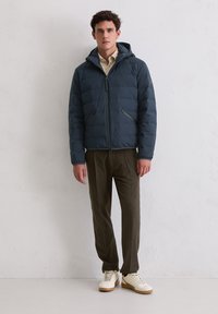 Marineblauw gewatteerd jack met kap, voorzien van een doorgestikt ontwerp, ritssluiting aan de voorkant en twee zijzakken. Gedragen over een beige shirt en olijfgroene broek.