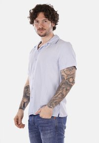 Camicia azzurra a maniche corte con design a bottoni, texture liscia e colletto. Indossata con jeans blu, con tatuaggi visibili sul braccio.