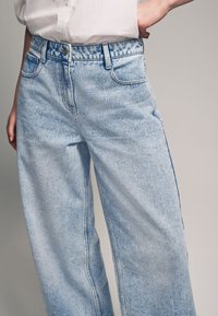 Jeans de mezclilla ligera de corte ancho en azul claro con un acabado desgastado, que cuentan con un cierre de botón, bolsillos frontales y detalles de costura visibles.