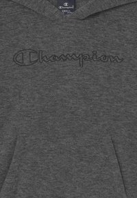 Grå hættetrøje i blødt, tekstureret stof med et syet "Champion"-logo i mørkegråt på tværs af brystet. Indeholder en frontlomme og hætte.