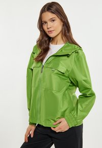 Grön huva jacka i lätt material, med dragkedja, två bröstfickor och elastiska mudd. Slät textur med en glänsande yta.