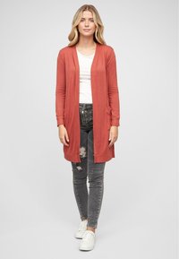 Lang, åpenfront cardigan i rustfarge, laget av myk stoff med ribbestrikkede mansjetter. Kombinert med svarte slitte jeans og hvite sneakers.