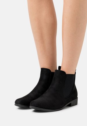 Bottes basses en daim noir avec panneaux latéraux élastiques, semelles en caoutchouc plates et un design épuré à bout arrondi, arborant un style minimaliste.