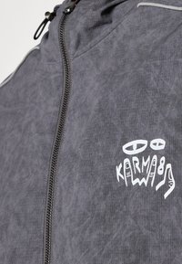 Gray zippered jacket with a textured fabric, featuring a white graphic print of hands and the word "KARMA" on the chest.
