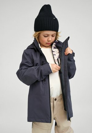 Wodoodporna ciemnoszara kurtka z wieloma kieszeniami zapinanymi na zamek oraz wzorzystą podszewką wewnętrzną, zestawiona z czarną prążkowaną czapką beanie.