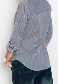 Camisa de algodón a rayas con mangas enrollables, colores azul marino y blanco, dobladillo curvado y textura suave. Combinada con jeans de mezclilla oscura.