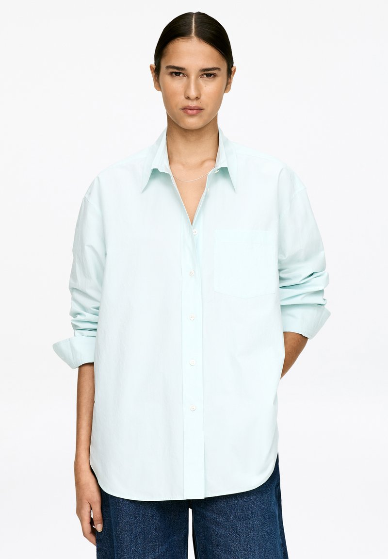 Chemise oversize vert menthe clair avec une seule poche poitrine et des manches retroussées, confectionnée en tissu de coton doux.