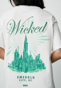 Valge puuvillane T-särk, millel on roheline graafika linnamaastikust, kus on kuplid ja teravad tornid. Tekst ütleb "Wicked" ja "Emerald City, OZ."