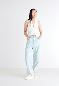 Hvitt halterneck-top med plisserte detaljer og innsnevret midje, parret med lyseblå vide jeans og hvite sneakers, satt mot en ensfarget bakgrunn.