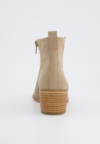 Bota de tobillo perforada beige con tacón de madera apilada y cierre de cremallera en la parte trasera. Presenta una punta redonda y una textura suave.