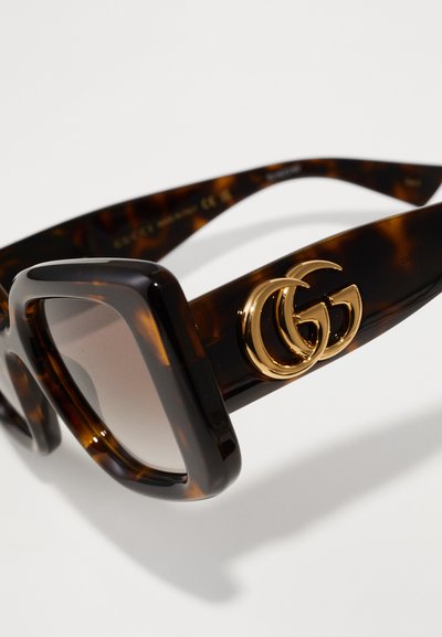 schildpadvormige zonnebril met een dik, vierkant montuur en gradient lenzen. Bevat een gouden GG-logo aan de zijkant, wat de luxe ontwerpelementen benadrukt.