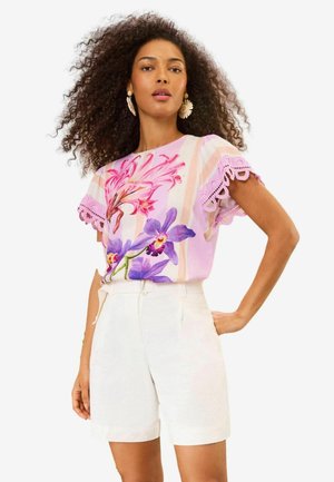 Göndör hajú nő, bézs rövidnadrágot és rózsaszín virágos felsőt csipkés ujjakkal visel, egyik kezét csípőjén tartja.