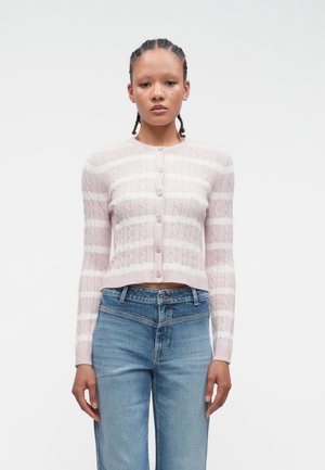 Femme aux cheveux tressés portant un cardigan boutonné à rayures rose et blanches et un jean bleu taille haute, se tenant contre un fond blanc.
