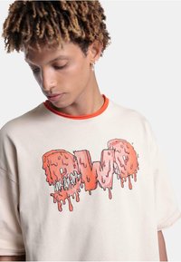 Beige oversized T-skjorte med oransjefarget hals, med et stort smeltende "OWO"-grafikk i rosa og røde nyanser. Myk bomullstekstur.