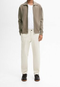 Lichtgewicht olijfgroene cardigan met ritssluiting over een wit T-shirt, gecombineerd met beige, recht gesneden broek en zwarte veterschoenen.