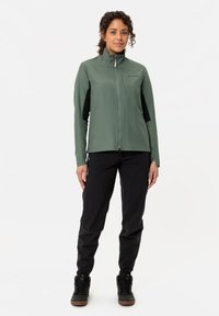 Giacca verde con zip e dettagli neri, colletto alto e materiale leggero abbinata a pantaloni neri e scarpe robuste scure in una posa eretta.