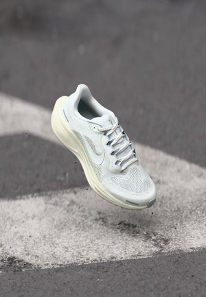 Chaussure de sport gris clair avec dessus en mesh, accents réfléchissants et semelle rembourrée, surélevée au-dessus d'une surface grise avec des marquages blancs.