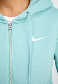 Gaiši zaļš kapuce ar rāvējslēdzēju, izgatavots no mīksta auduma, ar baltu Nike logo kreisajā krūtī un aukluakcentiem uz kapuces.