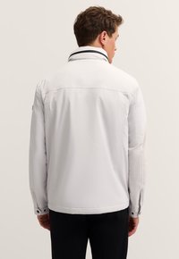 Mann in einer weißen Jacke mit hohem Kragen, der mit dem Rücken zur Kamera vor einem schlichten, hellen Hintergrund steht.