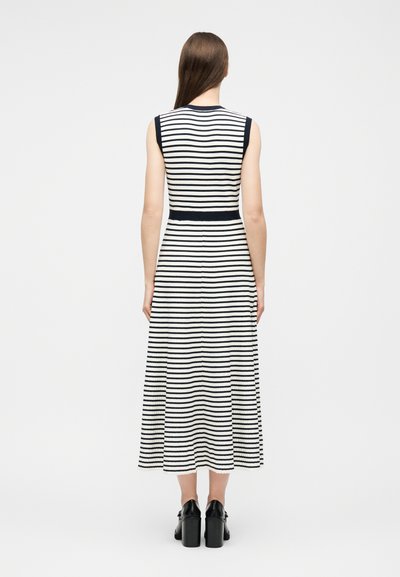 Femme aux longs cheveux bruns portant une robe midi sans manches à rayures horizontales noires et blanches et des chaussures à talons noires, debout devant un fond uni.