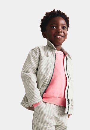 Veste à rayures grises claires et blanches avec une fermeture éclair à l'avant, associée à un sweat-shirt rose. Le mannequin sourit et porte un pantalon à rayures assorti.