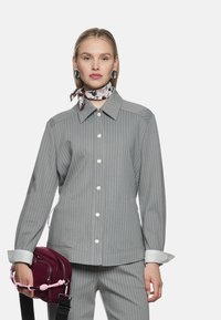 Graues Nadelstreifenhemd mit klassischem Kragen, weißen Knöpfen und kontrastierenden weißen Manschetten. Das Modell hält eine kleine burgunderrote Tasche mit rosa Griff.