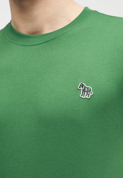 Πράσινο μπλουζάκι με στρογγυλή λαιμόκοψη, με ένα μικρό πολύχρωμο ριγέ πατς ζέβρας στο αριστερό στήθος, που φοριέται από ένα άτομο, με ορατό λαιμό και ώμους.