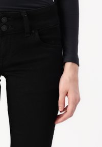 Rechte Hand entspannt an der Seite, trägt ein schwarzes Langarmoberteil und hochgeschnittene schwarze Jeans mit doppeltem Knopfverschluss.