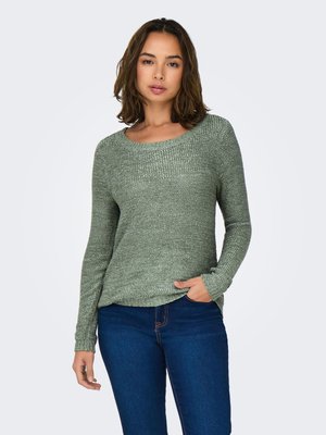 Femme aux cheveux bruns mi-longs portant un pull vert en maille et un jean bleu foncé, debout avec une main dans la poche, sur un fond blanc.