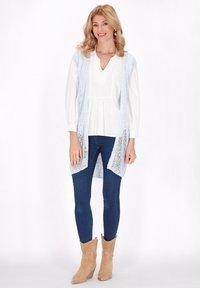 Un lungo gilet lavorato a maglia azzurro chiaro su una camicia bianca, abbinato a jeans blu scuro e stivaletti beige con dettagli perforati.