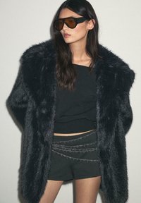 Kobieta w oversize'owym czarnym płaszczu z sztucznego futra, czarnym topie, czarnych szortach z srebrnym łańcuchem oraz dużych okularach przeciwsłonecznych w brązowym odcieniu.