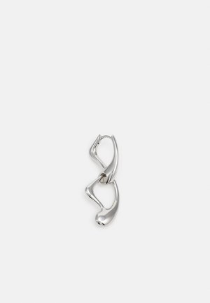 Boucles d'oreilles en forme de créoles abstraites ton argent avec des surfaces lisses et courbes, reliées par une petite charnière sur un fond gris clair.