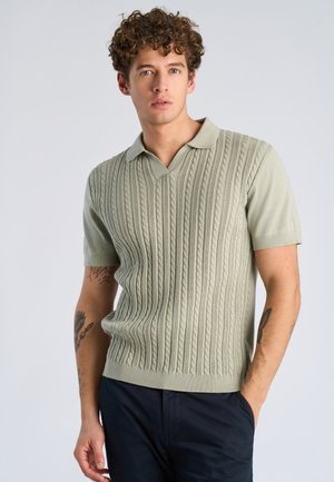 Młody mężczyzna z kręconymi włosami, ubrany w jasnografitowy sweter typu polo z krótkim rękawem o warkoczowym splocie oraz ciemne spodnie, stojący na jednolitym tle.