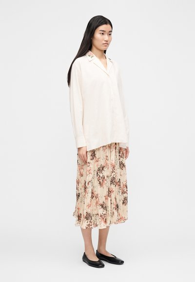 Mulher com cabelo longo e escuro, vestindo uma camisa creme com botões, saia plissada floral e sapatilhas pretas, de pé contra um fundo branco simples.