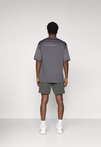 Camiseta atlética gris de manga corta y textura suave, combinada con pantalones cortos verdes. El fondo es simple y claro.