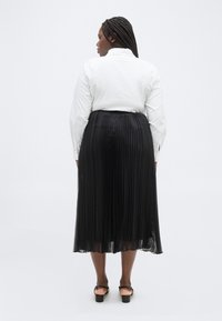 Γυναίκα με πλεξούδες, φοράει λευκή μακρυμάνικη μπλούζα, μαύρη πλισέ midi φούστα και μαύρα σανδάλια με τακούνι, στέκεται με την πλάτη προς το μέρος μας.