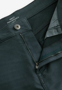 Pantaloni slim-fit de culoare verde închis, cu închidere cu fermoar și buton metalic. Materialul pare neted, având textură minimă și detalii de cusătură curate.