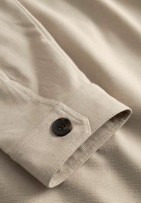 Poignet de chemise beige à manches longues avec un bouton sombre à quatre trous, montrant un tissu finement texturé et une couture détaillée.