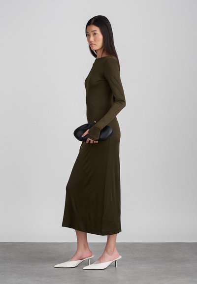 Vihreä oliivinvärinen mekko, jossa on pitkät hihat, polven pituinen helma ja ribatoitu pinta. Musta soikea clutch-laukku ja valkoiset kärkitossut täydentävät asun.