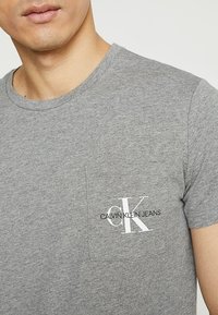 Camiseta de algodón gris con diseño de manga corta, cuello redondo y un bolsillo en el pecho izquierdo que presenta un logo impreso de "CK" y el texto "CALVIN KLEIN JEANS".