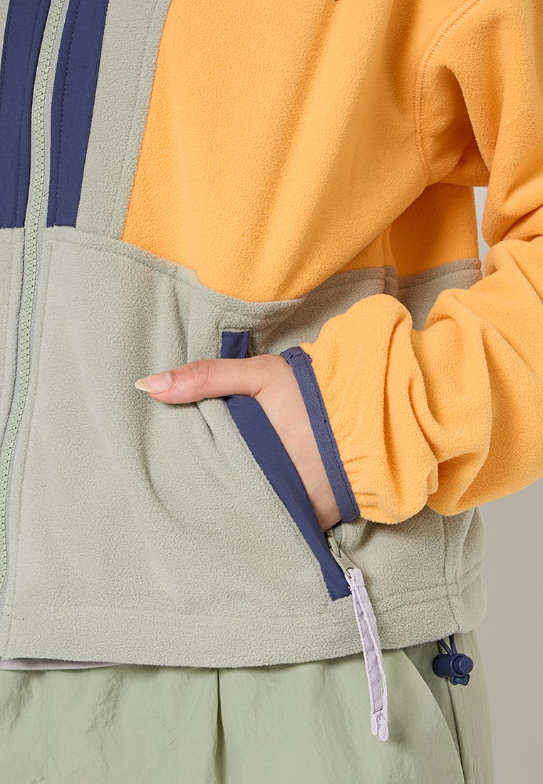 Mano en un bolsillo de chaqueta de forro polar gris y naranja con ribete y cremallera azul marino, usada con pantalones verde claro.