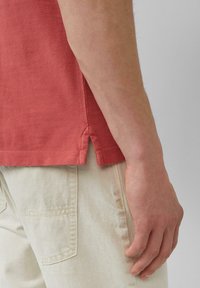 Kurzes rotes Hemd aus strukturiertem Stoff, mit seitlichen Schlitzen am Saum. Helle Hose mit sichtbarem Vordertasche.