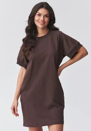 Uśmiechnięta kobieta o długich ciemnych włosach nosi luźną, krótką sukienkę w ciemnobrązowym kolorze, pozując z jedną ręką na biodrze na jasnym tle.