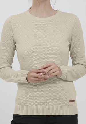 Beżowy sweter dzianinowy o fakturze, z długimi rękawami, noszony przez osobę z delikatnie złożonymi rękami na talii.