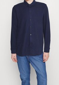 Hombre vestido con una camisa de botones de manga larga azul marino oscuro y jeans azules, de pie frente a un fondo claro y sencillo.