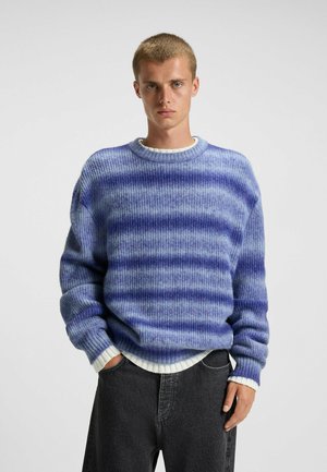 Blue and purple striped sweater made of soft knit material, featuring a crew neck and dropped shoulders, paired with gray jeans.