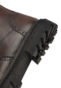Bottine en cuir marron avec des détails de brogue, présentant une semelle en caoutchouc noir texturé avec des crampons profonds pour une adhérence et une stabilité accrues.
