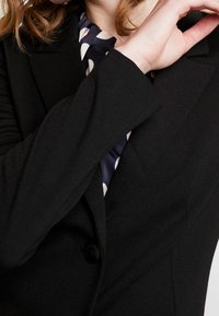 Svart blazer med en strukturerad design, enkel knappstängning och smala ärmar, lagd över en marinblå scarf med prickar.