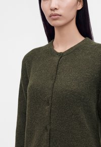 Femme portant un cardigan en maille duveteuse vert foncé, boutonné, avec un col rond sur un fond clair.