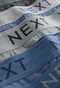 Sous-vêtements en tissu doux avec des bandes élastiques de couleurs grise et bleu foncé, arborant le logo "NEXT" en couture contrastée. Surfaces texturées visibles.